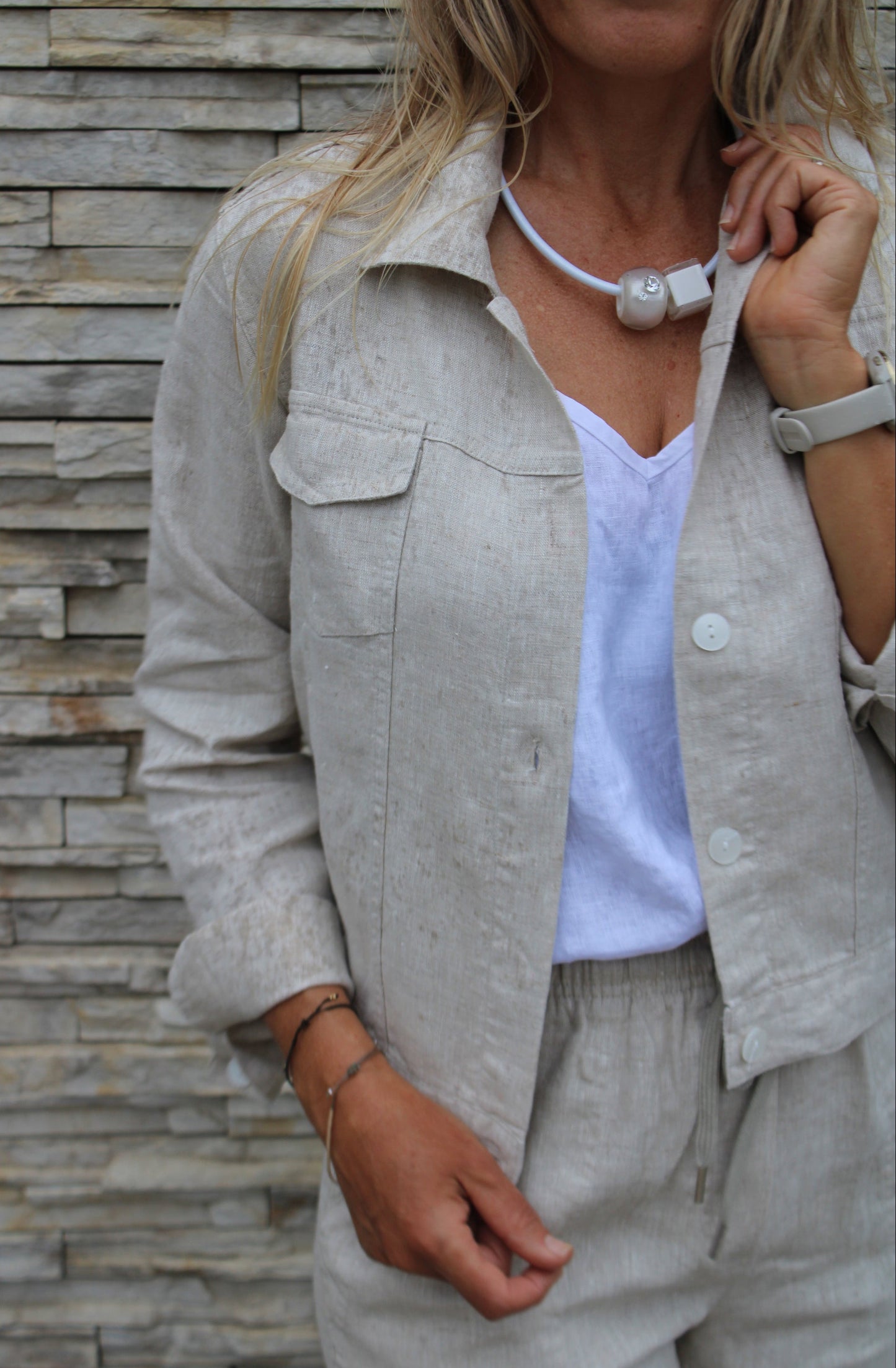 Person wearing a light natural jacket and white tank top against a stone wall.